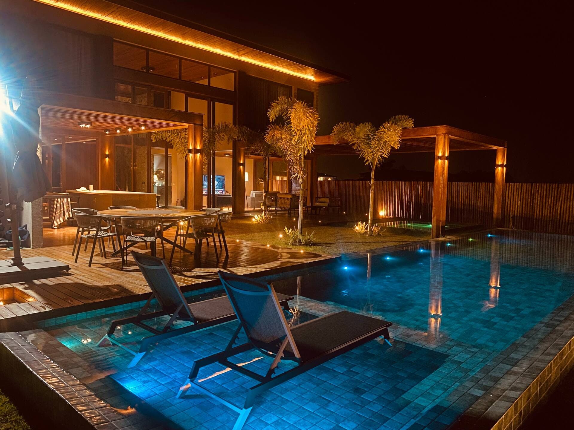 Deck com mesa de jantar e espreguiçadeiras dentro da piscina, à noite, com o beiral de madeira iluminado