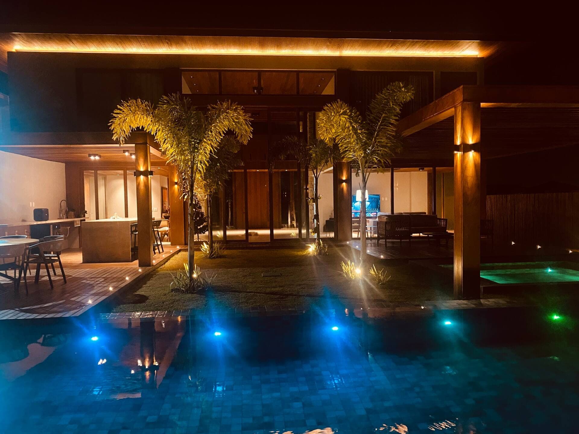 Piscina com luzes azuis à noite, palmeiras e a área gourmet ao fundo