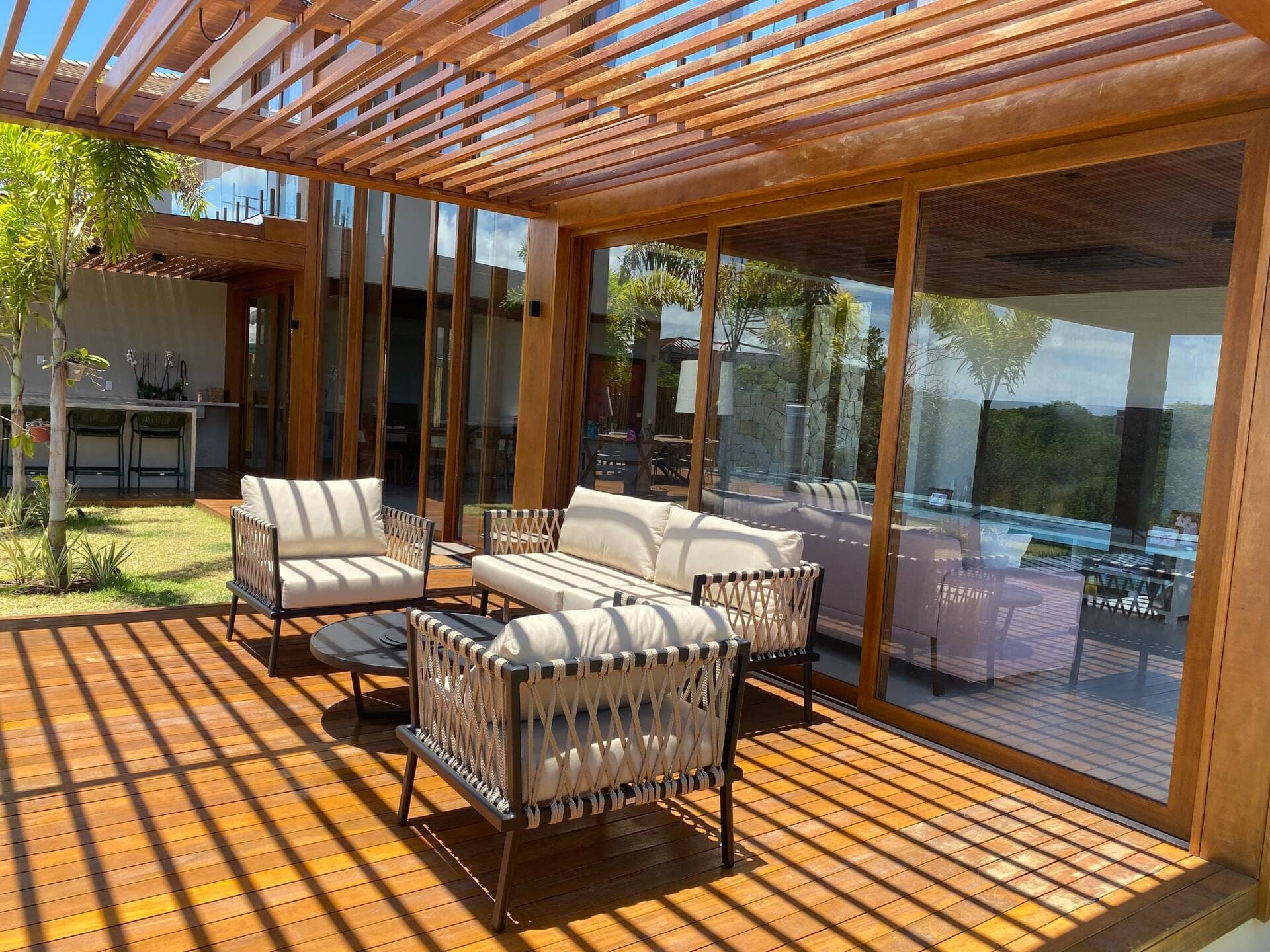 Pergolado de madeira com sofás e poltronas sobre o deck, aberto para o jardim