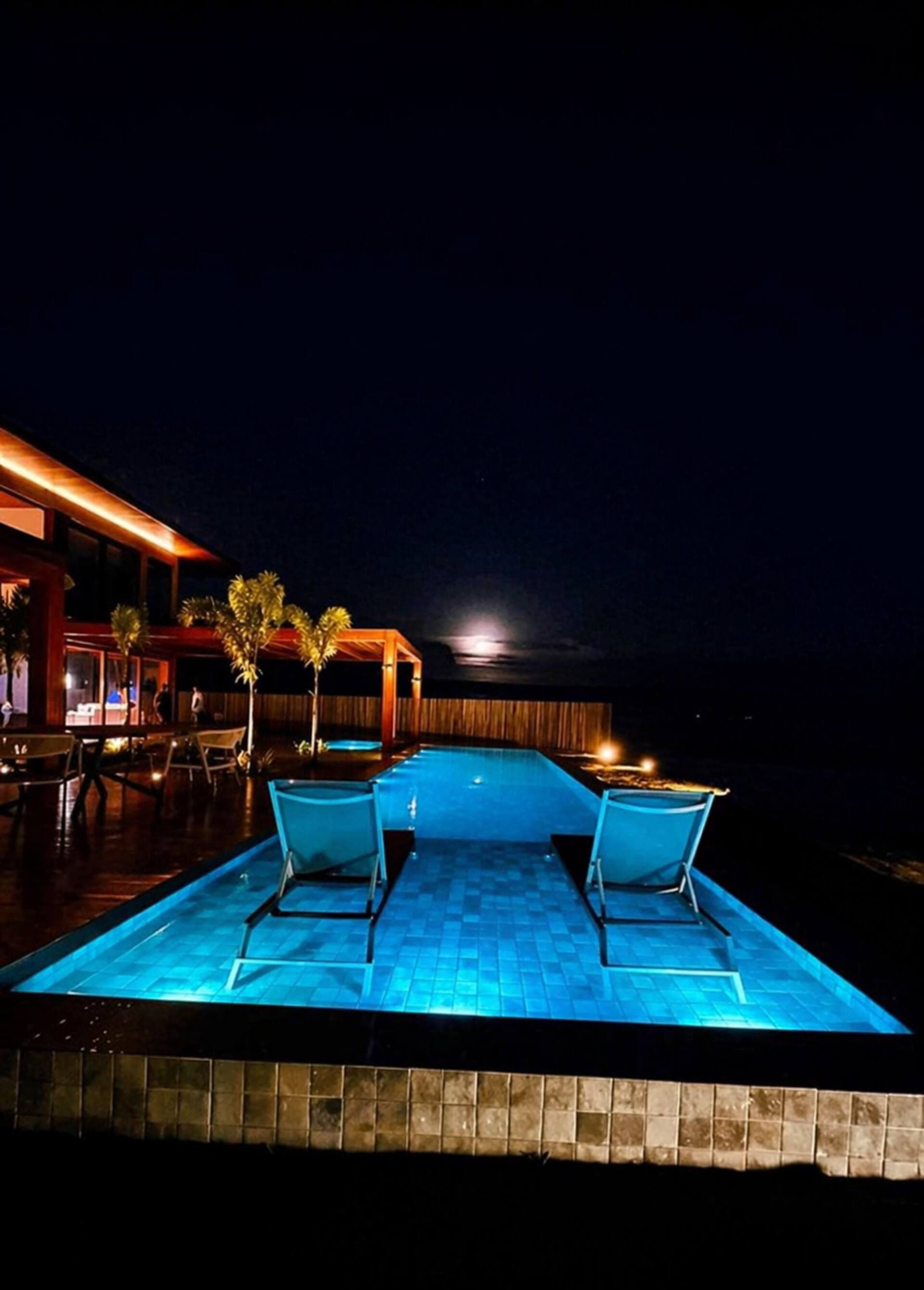 Piscina iluminada à noite, com duas espreguiçadeiras, e o beiral aceso ao fundo