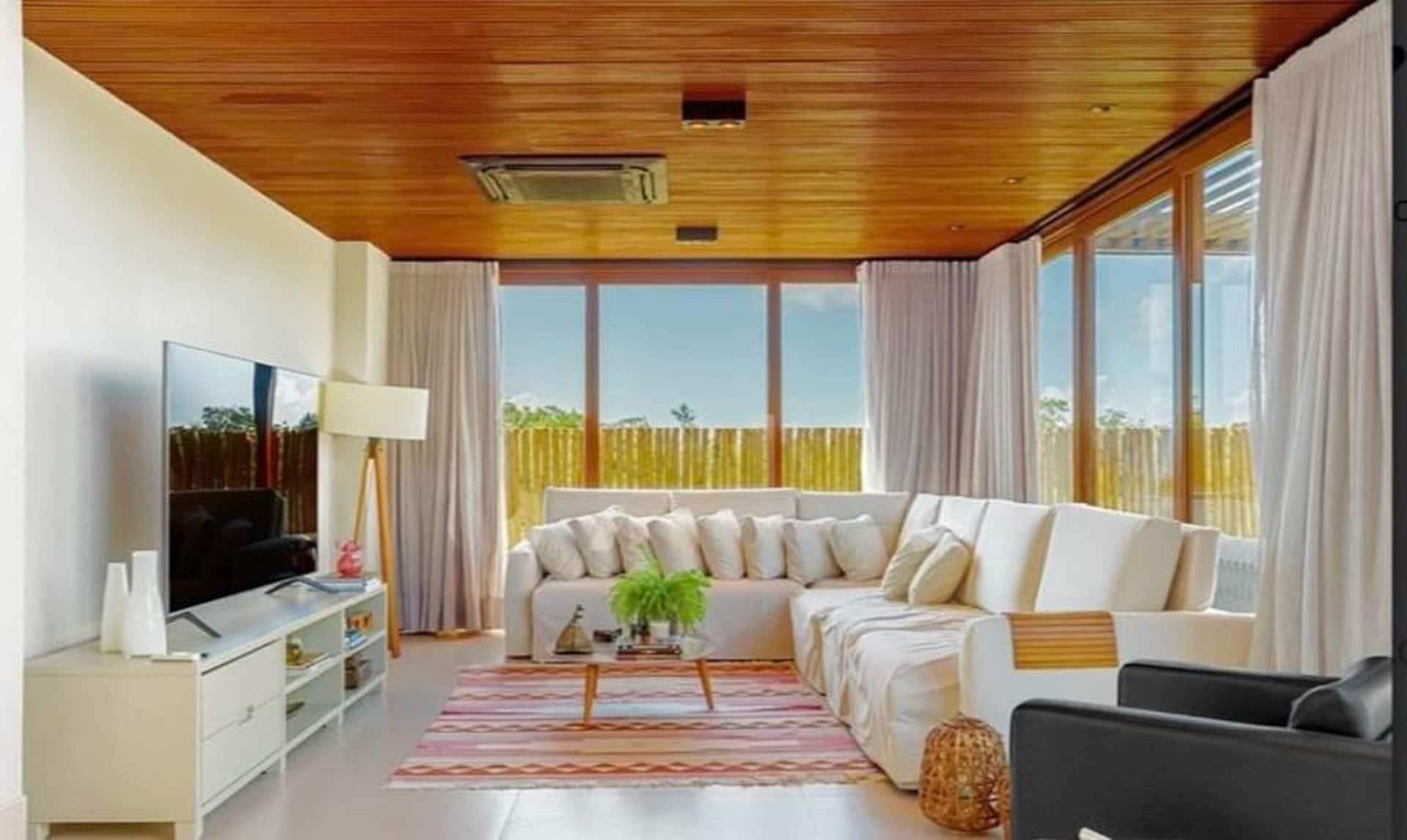 Sala de estar com sofá em L, TV e cortinas, aberta para o jardim